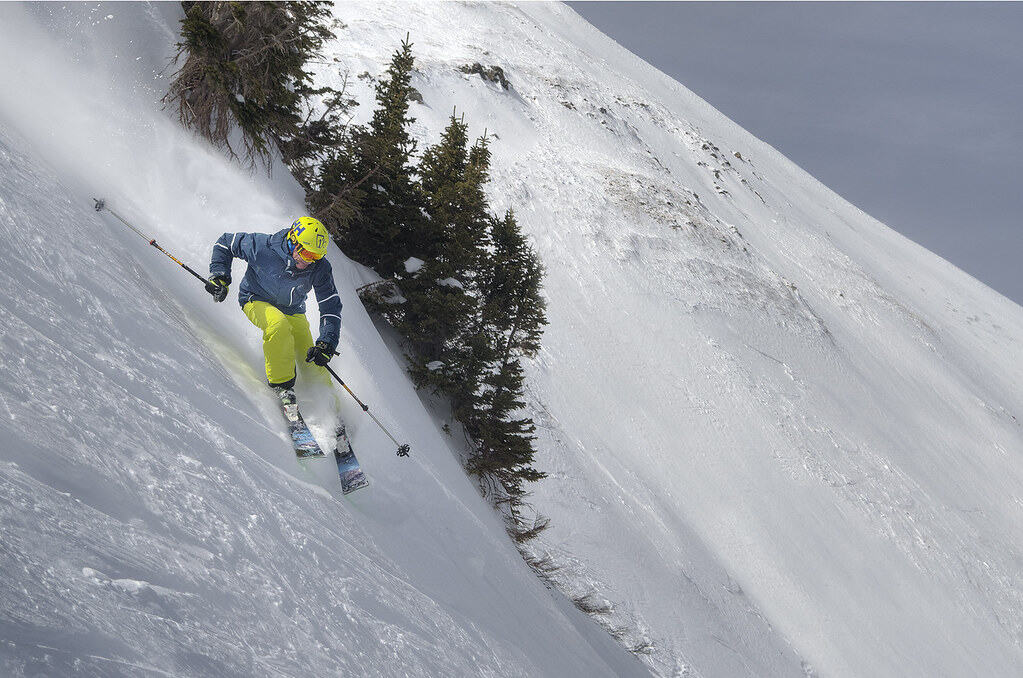 Man skiing down a steep slope with the mountainside and trees in the background